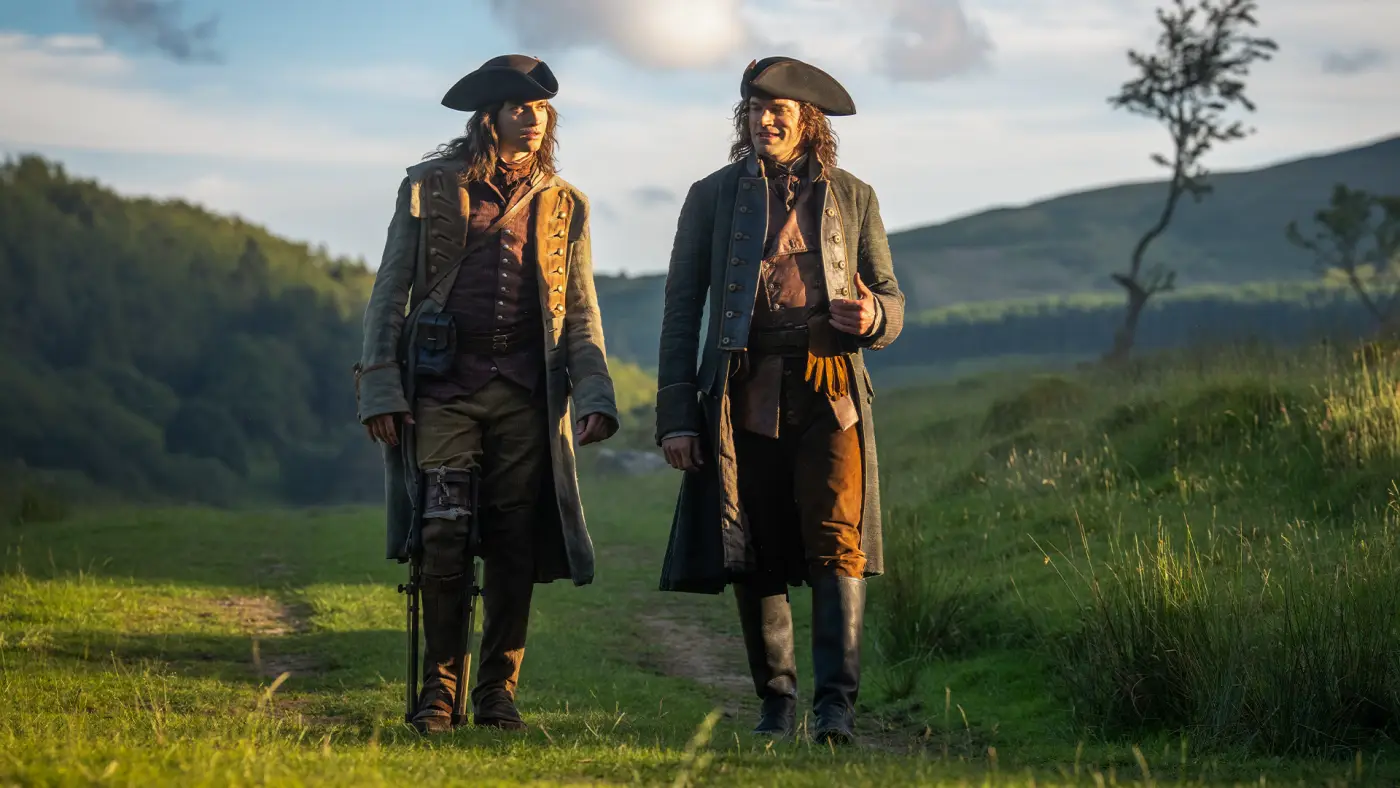 Two brothers in colonial coats walk side by side across a green hillside.